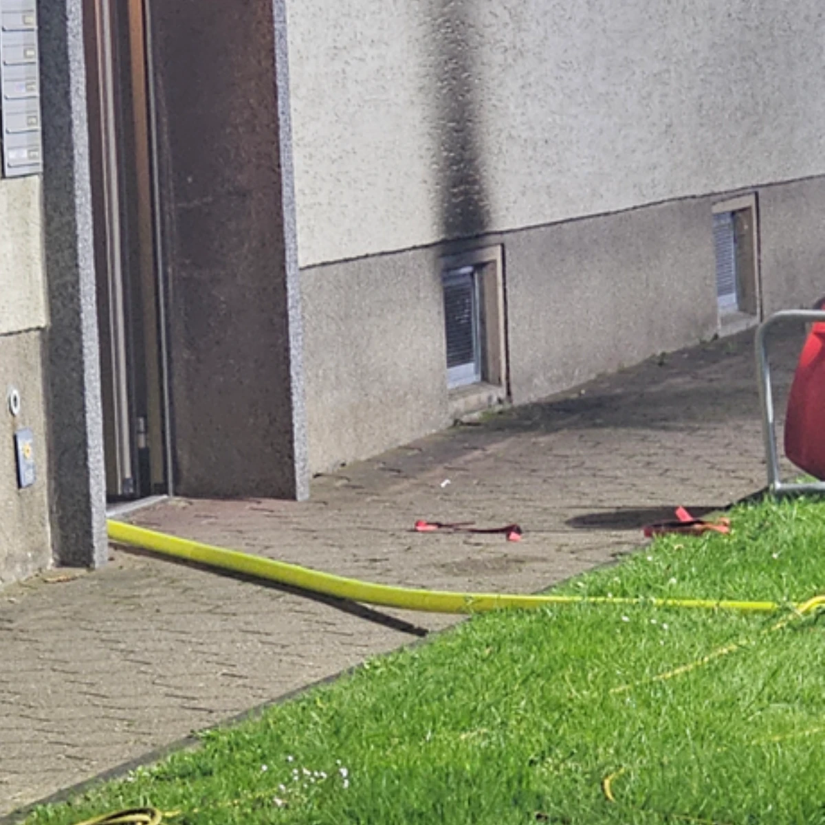 Außenfassade eines Wohngebäudes Außenfassade eines Wohngebäudes mit deutlicher Rußspur nach einem Brand; Feuerwehrschlauch und Werkzeug liegen im Gartenbereich.