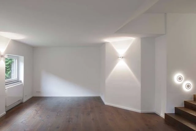 Renovierungsarbeiten innen Maler Scholz 1 Renovierter Wohnraum mit weiß gestrichenen Wänden, moderner Wandbeleuchtung und dunklem Holzfußboden.