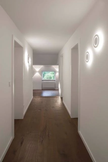 Renovierungsarbeiten innen Maler Scholz 2 Heller, frisch renovierter Flur mit weißen Wänden, moderner Wandbeleuchtung und Blick in ein Zimmer mit Fenster.