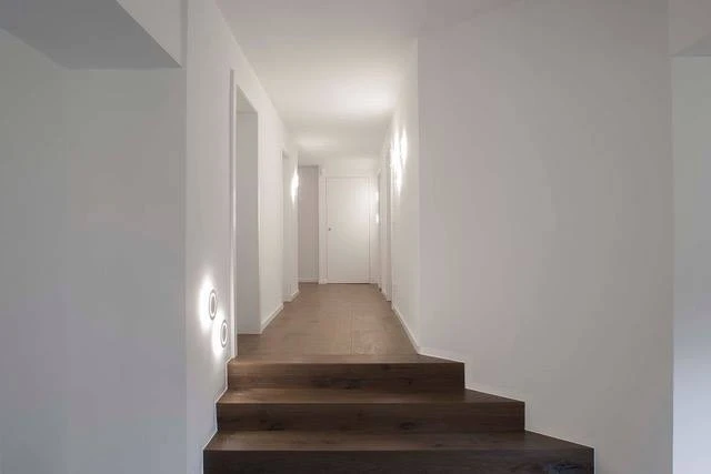 Renovierungsarbeiten innen Maler Scholz 4 Langer Flur mit weißen Wänden und einer dunklen Holztreppe, minimalistische Wandbeleuchtung an den Seiten.