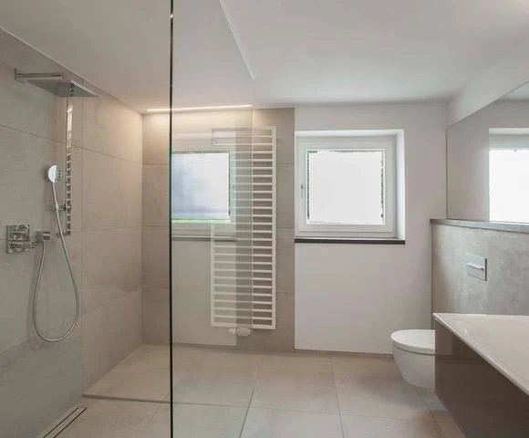 Renovierungsarbeiten innen Maler Scholz 5 Modernes Badezimmer mit bodengleicher Glasdusche, Handtuchheizkörper und wandhängendem WC, helle neutrale Farben.